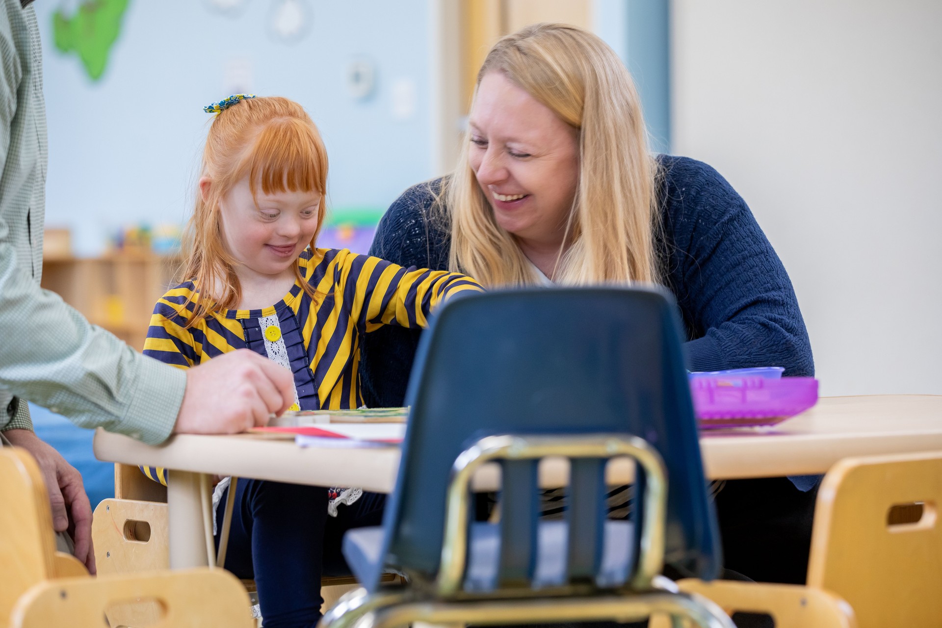 A cute little girl with red hair and Down Syndrome plays at a small table in a classroom while her mom smiles at her.